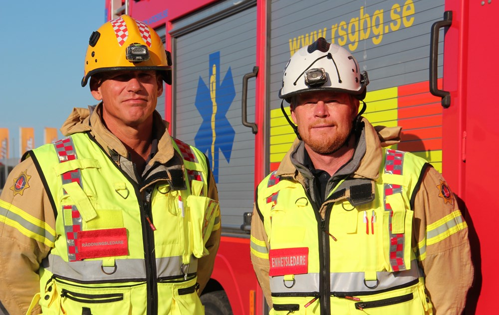 Två brandpersonal i gula larmställ och gula västar. En med vit och en med röd hjälm